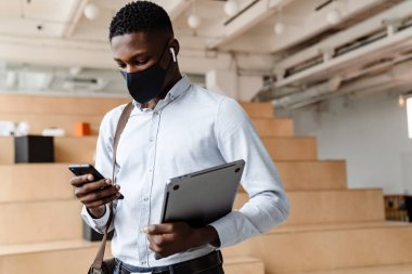 Sınıfta duran, dizüstü bilgisayarını tutan, cep telefonu kullanan, tıbbi maske takan kendinden emin Afrikalı öğrenci.