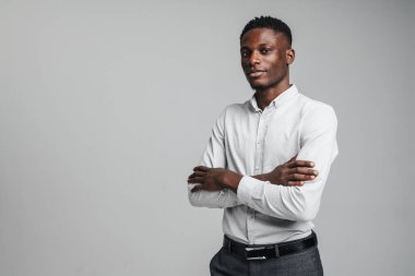 Zeki beyaz gömlek giyen, kendine güvenen genç Afrikalı adam gri arka planda tek başına duruyor, kolları bağlı.