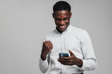 Yakışıklı, heyecanlı, Afrikalı iş adamı. Gri arka planda izole edilmiş cep telefonu kullanıyor. Beyaz resmi gömlek giyiyor ve başarısını kutluyor.