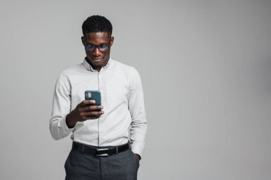 Gri arka planda izole edilmiş beyaz resmi tişört giyen yakışıklı, kendine güvenen Afrikalı bir iş adamı cep telefonu kullanıyor.