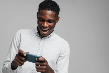 Yakışıklı, heyecanlı, Afrikalı iş adamı cep telefonuyla oyun oynuyor. Gri arka planda izole edilmiş, beyaz resmi tişört giyiyor.