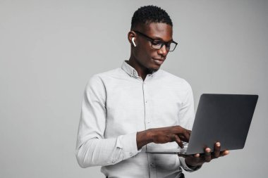 Gri arka planda izole edilmiş dizüstü bilgisayar kullanan, kablosuz kulaklıklı, yakışıklı, Afrikalı bir iş adamı.