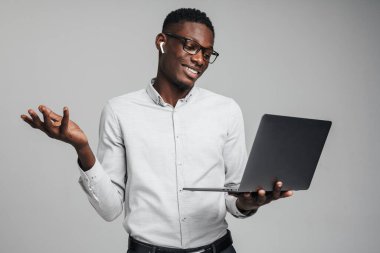 Yakışıklı, gülümseyen Afrikalı iş adamı dizüstü bilgisayarını gri arka planda tutuyor ve video aracılığıyla iş arkadaşlarıyla konuşuyor.