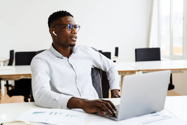 Attractive young dark-skinned businessman wearing formal clothing and eyeglasses conducting business negotiations with client through video-chat on laptop computer, using earphones