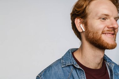 Kablosuz kulaklıklı neşeli kızıl saçlı adam beyaz arka planda izole edilmiş kameraya gülümsüyor.