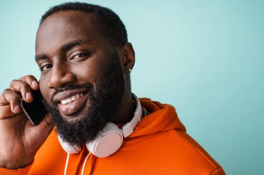 Gülümseyen Afrikalı genç adam mavi arka planda izole edilmiş cep telefonuyla konuşuyor.