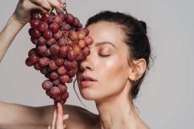 Gri arka planda izole edilmiş üzümlerle poz veren güzel yarı çıplak kadın.