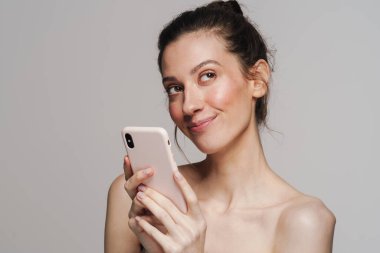 Gri arka planda izole edilmiş cep telefonu kullanırken gülümseyen mutlu yarı çıplak kadın.