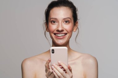 Gri arka planda izole edilmiş cep telefonu kullanırken gülümseyen mutlu yarı çıplak kadın.