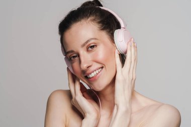 Gri arka planda izole edilmiş kulaklık takıp gülümseyen mutlu yarı çıplak kadın.