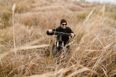 Tıraşsız, güneş gözlüklü, bisikletli bir adam sahada yürüyor.