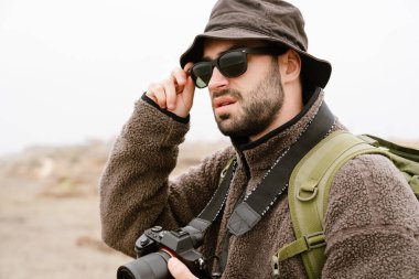 Güneş gözlüklü yakışıklı fotoğrafçı dışarıda dijital kamerayla yürüyor.