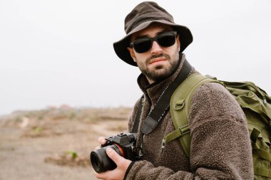 Güneş gözlüklü yakışıklı fotoğrafçı dışarıda dijital kamerayla yürüyor.