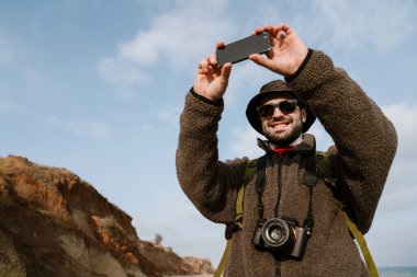 Plajda cep telefonuyla selfie çekerken gülümseyen mutlu traşsız adam.