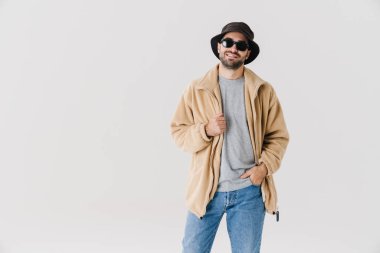 Güneş gözlüklü neşeli yakışıklı adam gülümsüyor ve beyaz arka planda izole edilmiş kameraya bakıyor.