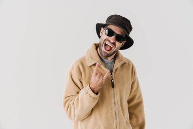 İzole edilmiş beyaz arka planda bej ceket, kep ve güneş gözlüğü takan mutlu bir gencin portresi jesti bozuyor.