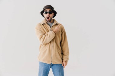 İzole edilmiş beyaz arka planda bej ceket, şapka ve güneş gözlüğü takan mutlu bir gencin portresi.