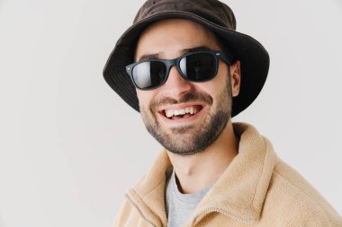 İzole edilmiş beyaz arka planda bej ceket, şapka ve güneş gözlüğü takan mutlu bir gencin portresi.