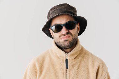 Güneş gözlüklü hoşnutsuz yakışıklı adam somurtuyor ve beyaz arka planda izole edilmiş kameraya bakıyor.