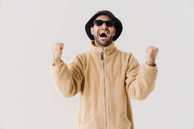 İzole edilmiş beyaz arka planda bej ceket, şapka ve güneş gözlüğü takan mutlu bir gencin portresi başarıyı kutluyor.