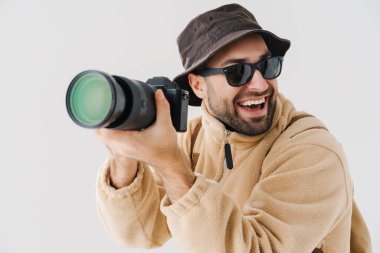 Güneş gözlüklü heyecanlı fotoğrafçı beyaz arka planda izole edilmiş dijital kamerayla fotoğraf çekiyor.