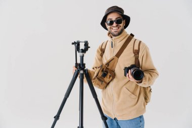 Beyaz zemin üzerinde izole edilmiş dijital kamera ve tripodla poz veren neşeli genç fotoğrafçı
