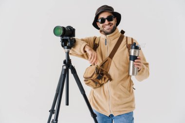 Beyaz arka planda izole edilmiş dijital kamerayla poz verirken çay içen neşeli genç fotoğrafçı