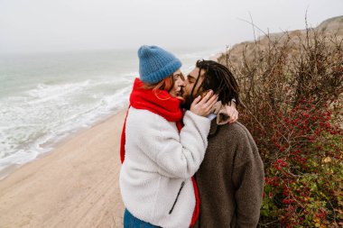 Sahilde kucaklaşan, kalın giysiler giyen, öpüşen çok ırklı bir çift.