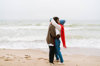Çok ırklı, güzel bir çift plajda sarılıyor, kalın giysiler giyiyor.
