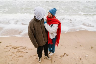 Çok ırklı, güzel bir çift plajda sarılıyor, kalın giysiler giyiyor.
