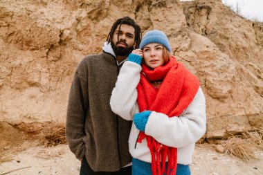 Çekici, soğuk, etnik çeşitlilikte bir çift sahilde yürüyorlar, kalın giysiler giyiyorlar.