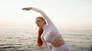 Ciddi bir kızıl saçlı kadın beyaz spor takım elbise giyiyor ve sabah deniz kenarında spor egzersizleri yapıyor.