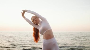 Beyaz spor kıyafeti giyen genç kızıl saçlı bir sporcu sabah deniz kenarında spor egzersizleri yapıyor.