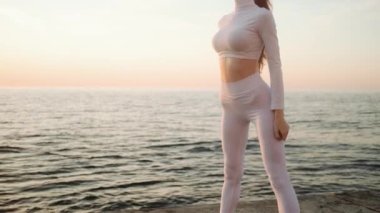 Yoğunlaşmış genç, kızıl saçlı, beyaz spor takım elbiseli bir kadın sporcu sabah deniz kenarında çömelme hareketleri yapıyor.