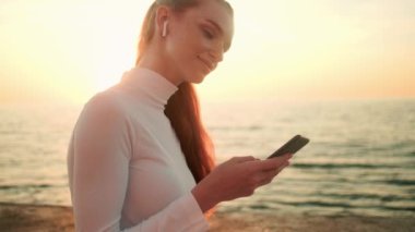 Beyaz spor elbise giyen genç bir kadının yan görüntüsü sabah deniz kenarında oturan akıllı telefonunu kullanıyor.