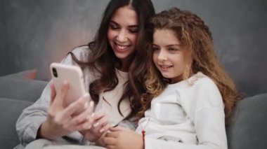 Gülümseyen anne ve kızı evdeki kanepede otururken akıllı telefon kullanıyorlar.