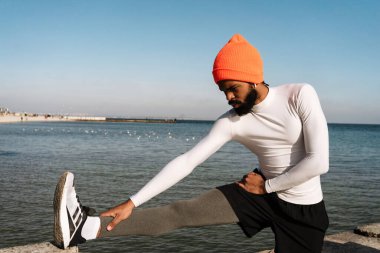 Atletik Afro-Amerikan kulaklıklı sporcu plajda çalışırken egzersiz yapıyor.