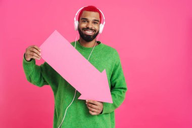 Heyecanlı genç Afro-Amerikalı 20 'li yaşlarda sokak kıyafetleri içinde pembe arka planda izole edilmiş, kulaklık takmış, okla işaret ediyor.