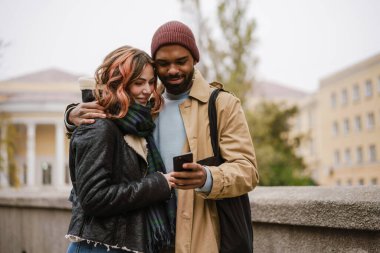 Çok uluslu gülümseyen çift şehir caddesinde sarılıp cep telefonu kullanıyor.
