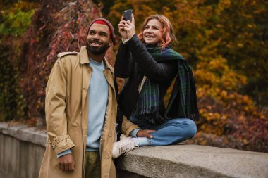 Çok uluslu mutlu çift gülümsüyor ve şehir caddesinde cep telefonuyla fotoğraf çekiyor.