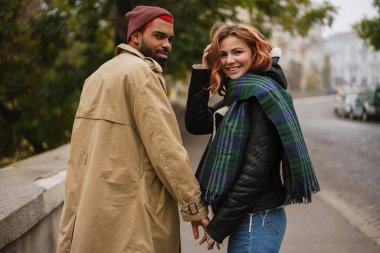 Çok uluslu mutlu çift el ele tutuşuyor ve caddede yürürken gülümsüyor.