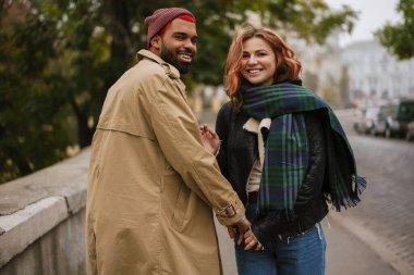 Çok uluslu mutlu çift el ele tutuşuyor ve caddede yürürken gülümsüyor.