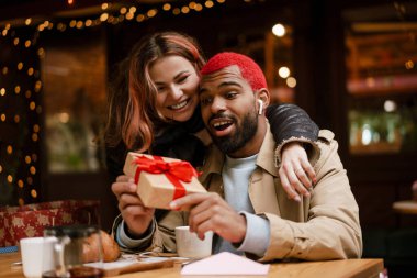 Çok uluslu mutlu çift gülümsüyor ve açık hava kafede hediye kutusunu tutuyorlar.