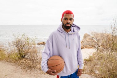 Kulaklıklı sakallı Afrikalı Amerikalı adam plajda basketbol oynuyor.