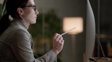 Gözlük takan ciddi bir iş kadınının yan görüşü ofiste elinde kalem tutarken bilgisayarla çalışmasıdır.