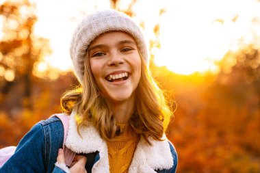 Örgü şapkalı gülen kız sonbahar parkında yürüyor, sırt çantası taşıyor.