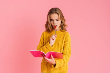 Pembe arka planda izole edilmiş egzersiz kitabıyla poz verirken kafası karışmış güzel sarışın kız.