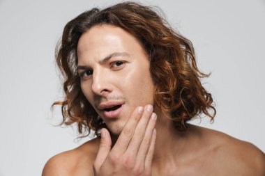 Kafası karışmış tişörtsüz adam yüzüne dokunuyor ve beyaz arka planda izole edilmiş kameraya bakıyor.