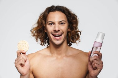 Kozmetik sünger ve temizlik köpüğüyle poz veren keyifli gömleksiz adam beyaz arka planda izole edilmiş.