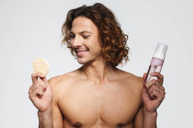 Beyaz arka planda izole edilmiş kozmetik sünger ve temizlik köpüğüyle poz veren mutlu üstsüz adam.